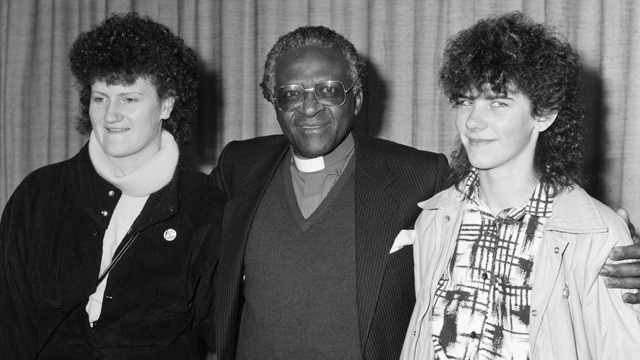 Bishop Desmond Tutu at London Airport, en route to Oslo to collect his Nobel prize, flanked by Irish women, Mary Manning (left) and Karen Gearon. 8 December 1984.
